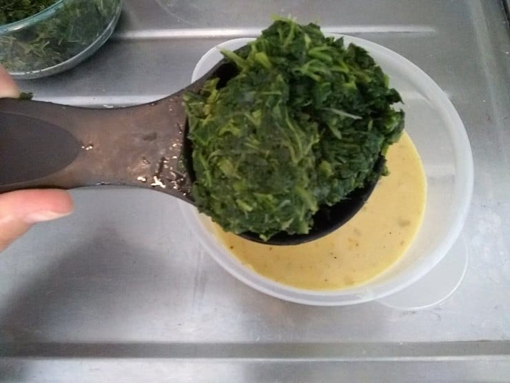 A spoonful of cooked spinach held over a measuring cup full of cheese sauce 