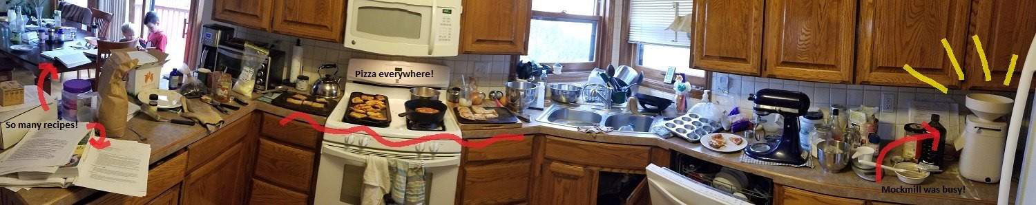 Counters, sink and stovetop covered with pizza ingredients, recipes, mixers, the mockmill grain mill and finished pizza.
