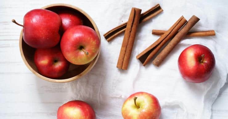 Apples stacked in a bowl. Whole cinnamon sticks and individual apples are laying next to the bowl.
