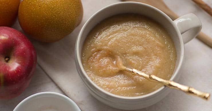 Homemade Instant Pot applesauce in a white ceramic bowl with a golden spoon surrounded by several whole apples of different varieties.